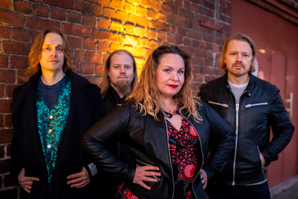 Kolme miestä ja nainen seisovat punatiiliseinän edessä ja katsovat kameraan.