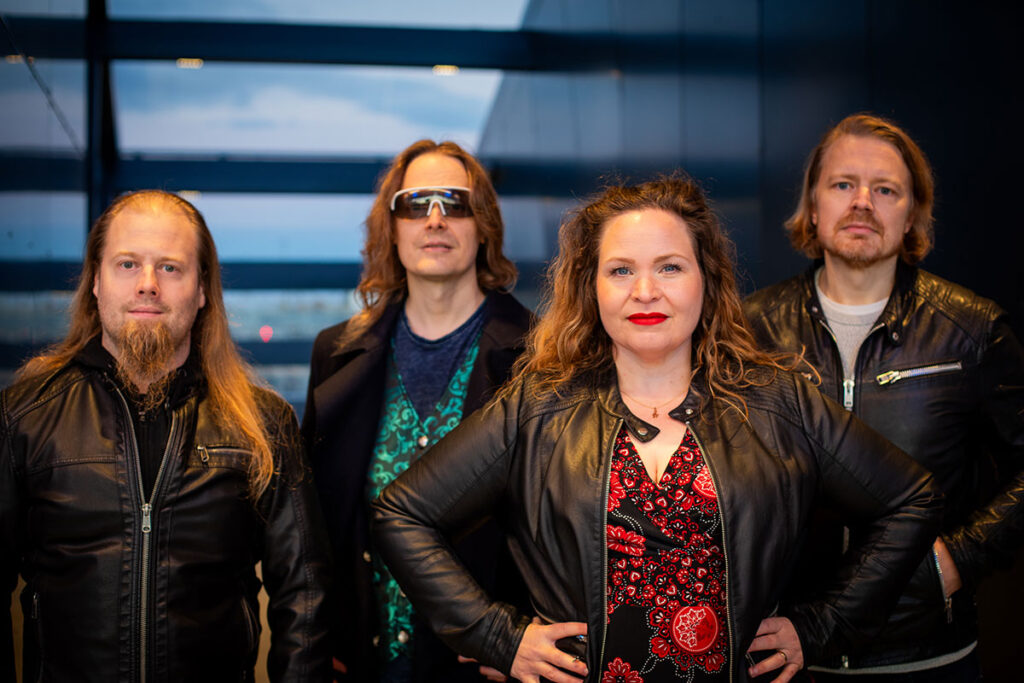 Nainen seisoo kolmen miehen edessä ja kaikki katsovat kameraan.